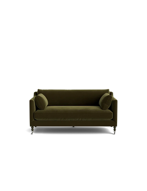 A Fabienne Sofa in green velvet, complete with two cylindrical cushions, set against a plain black background. The sofa boasts a modern design with clean lines and is supported by small casters.