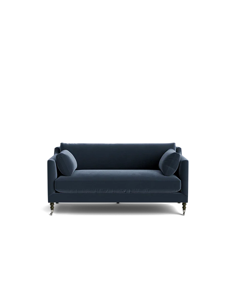 A Fabienne Sofa in dark blue velvet, featuring two matching round cushions, is set against a black background. The sofa showcases slim armrests and wooden legs with wheels.