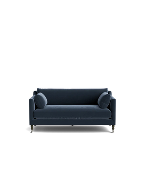 A Fabienne Sofa in dark blue velvet, featuring two matching round cushions, is set against a black background. The sofa showcases slim armrests and wooden legs with wheels.