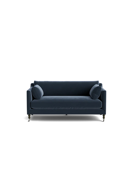 A Fabienne Sofa in dark blue velvet, featuring two matching round cushions, is set against a black background. The sofa showcases slim armrests and wooden legs with wheels.