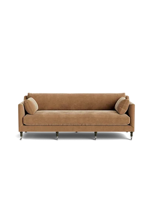The Fabienne Sofa, upholstered in beige velvet and accompanied by two matching cylindrical pillows, rests on six wooden legs set against a black background, showcasing its simple, modern design with clean lines.