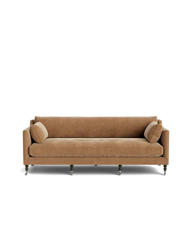 The Fabienne Sofa, upholstered in beige velvet and accompanied by two matching cylindrical pillows, rests on six wooden legs set against a black background, showcasing its simple, modern design with clean lines.