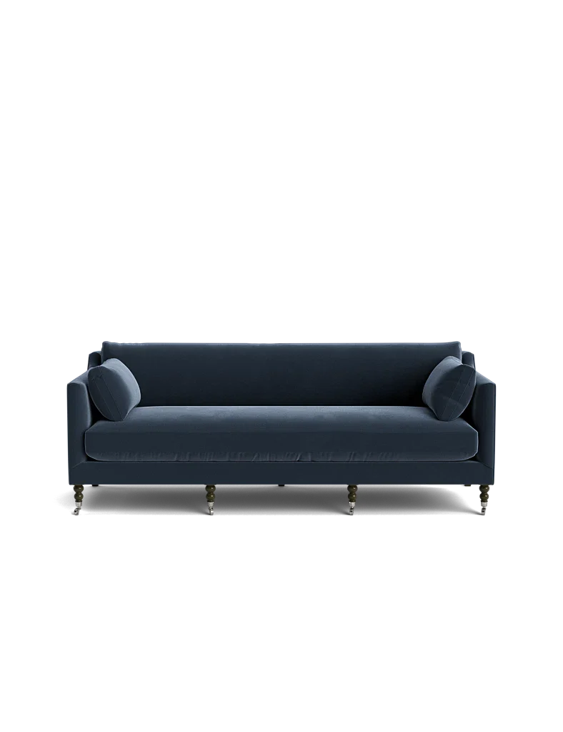The Fabienne Sofa, in a dark blue hue, features a modern design with clean lines and cylindrical cushions set against a black backdrop. Its four visible legs add an elegant touch to its simple yet stylish appearance.