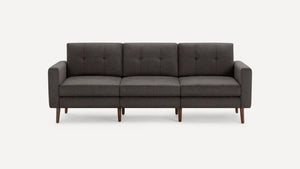 The Nomad Fabric Sofa, a modern three-seater in dark gray with square cushions and wooden legs, is showcased against a plain white background. It boasts clean lines and a minimalist design.