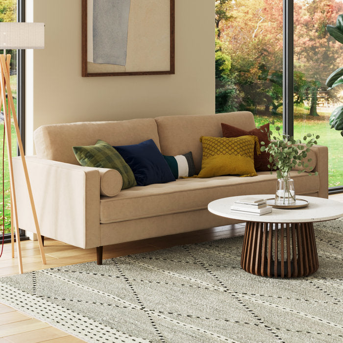 A cozy living room featuring the Geo 84'' Upholstered Sofa in a beige shade, adorned with colorful pillows; a round wooden coffee table topped with a vase of greenery; and a large window showcasing a view of trees. A floor lamp and a framed picture complete the decor.