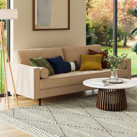 A cozy living room featuring the Geo 84'' Upholstered Sofa in a beige shade, adorned with colorful pillows; a round wooden coffee table topped with a vase of greenery; and a large window showcasing a view of trees. A floor lamp and a framed picture complete the decor.