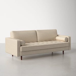 The Geo 84'' Upholstered Sofa in beige, featuring tufted cushions, cylindrical arm pillows, and wooden legs, is showcased on a light gray floor with a white background.