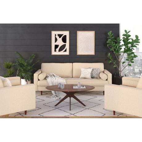 A contemporary living room features a Geo 84'' upholstered sofa in a neutral beige tone, accompanied by a round wooden coffee table and abstract art adorning a dark shiplap wall. Two potted plants flank the sofa, while large windows provide an expansive city view. A geometric rug introduces texture to the setting.