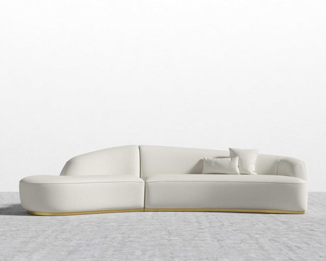 The Reya Curved Sectional is a sleek, modern sofa with soft lines and a minimalist design, featuring three matching cushions. It is displayed against a plain white wall on a light gray textured floor.