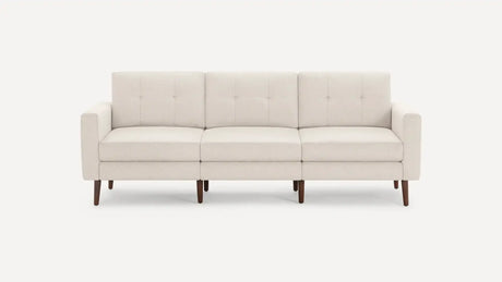 The Nomad Fabric Sofa is a light gray, three-seater with tufted back cushions, wooden legs, and a minimalist design, set against a plain white background.