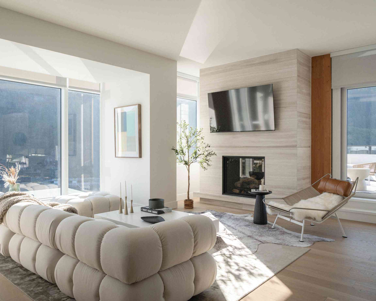 A contemporary living room featuring a spacious Belia Modular Sectional, a wall-mounted TV positioned above a cozy fireplace, and an elegant leather chair. Sunlight pours in through tall windows, brightening a small tree and minimalist decor. The floor boasts light wood accented by a patterned rug.