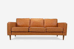 The Albany Sofa, featuring a mid-century modern design in warm tan vegan leather, has three cushioned seats and wooden legs, set against a plain white background.