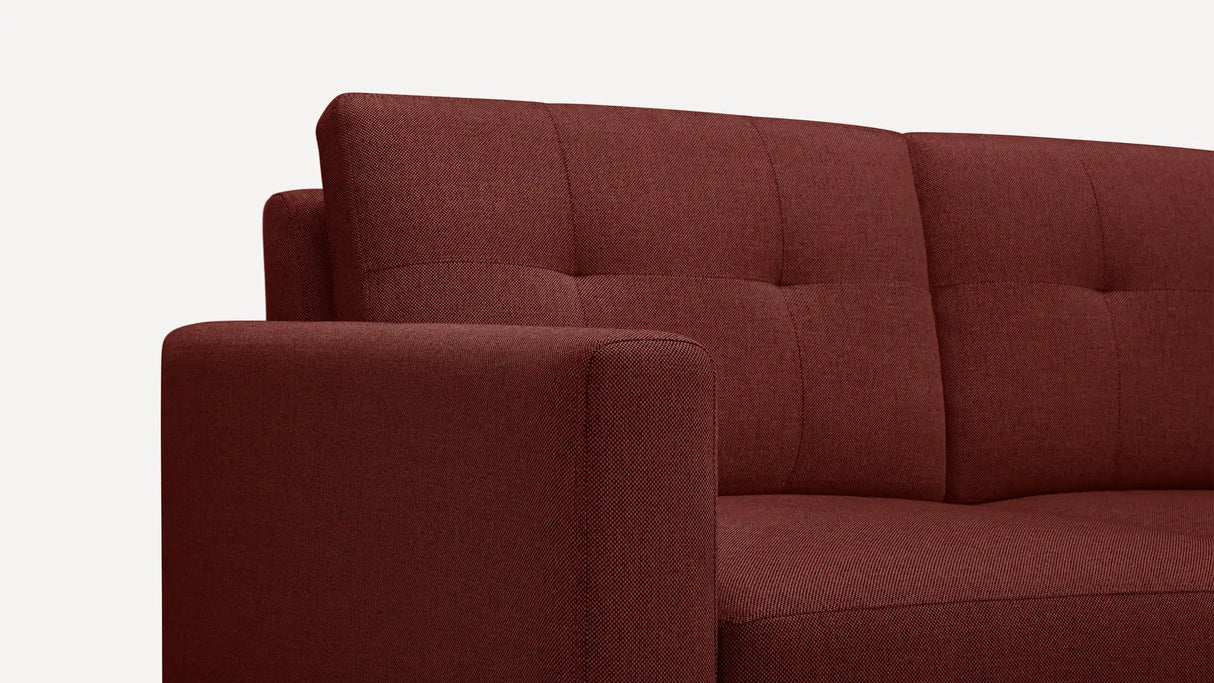 A detailed view of the Nomad Fabric Sofa reveals its plush, softly cushioned texture. The red upholstery has a grid-like design due to delicate stitching, with the armrest and backrest standing out against a plain white background.