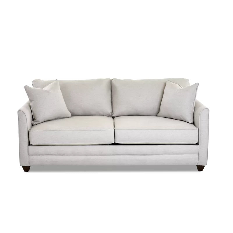 The Kodie 76.5" Upholstered Sleeper Sofa is a light gray piece featuring two seat cushions and matching back cushions, complemented by two square pillows on each side. It boasts a clean, modern design with slightly flared arms and rests on short dark wooden legs.