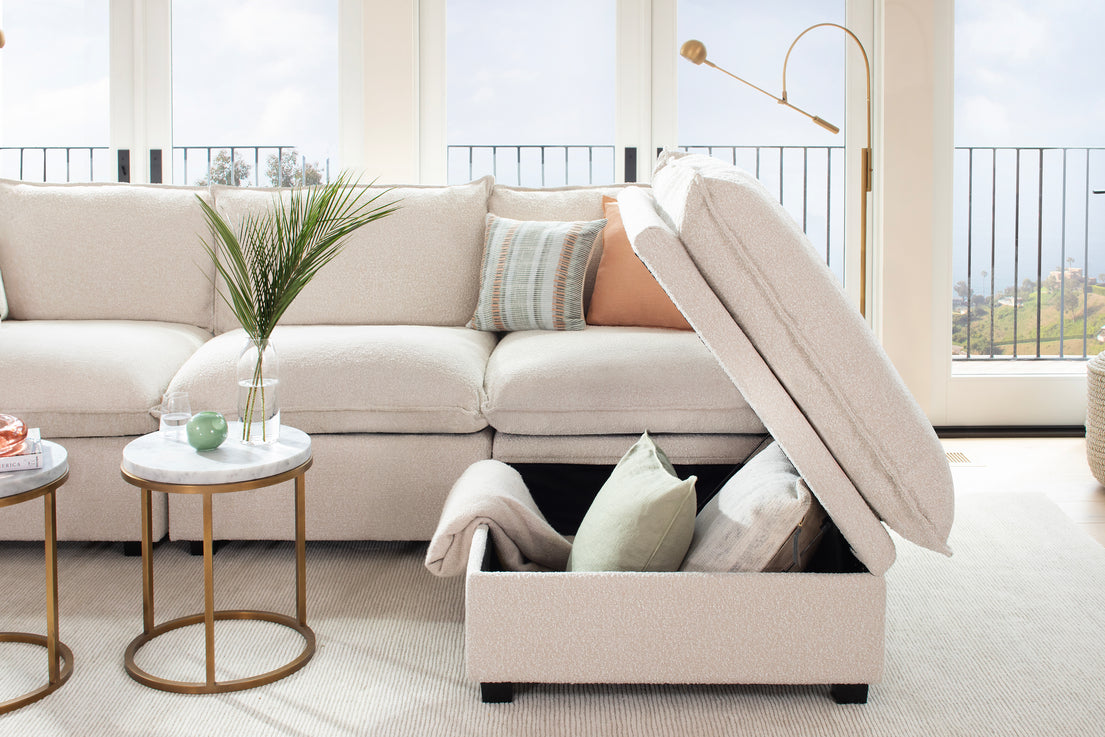 The generous living room features a Kova L Shape with Ottoman sectional sofa in light beige, complete with storage and decorated with patterned and solid pillows. Beside it are two round side tables adorned with plants and decorative items, all set against large windows showcasing a scenic landscape view.