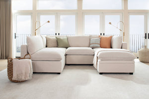 A cozy living room features a Kova L Shape with Ottoman sectional sofa adorned with pastel pillows in green, peach, and striped patterns. Behind it are large windows allowing natural light, with elegant floor lamps on each side. A wicker basket with a blanket sits nearby.