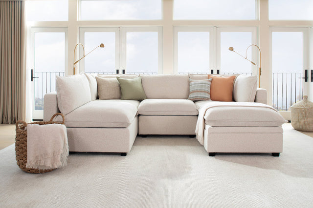 A cozy living room features a Kova L Shape with Ottoman sectional sofa adorned with pastel pillows in green, peach, and striped patterns. Behind it are large windows allowing natural light, with elegant floor lamps on each side. A wicker basket with a blanket sits nearby.