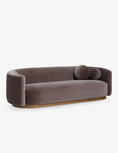 Introducing the Lowry Sofa: a contemporary piece in dark gray velvet featuring a rounded design with two cylindrical cushions adorning one side. It boasts smooth curves and a wooden base, beautifully displayed against a plain white background.