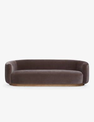 The Lowry Sofa is a stylish piece with its dark gray velvet upholstery, rounded arms, and low backrest. Set against a plain white background, it boasts a sleek, minimalist design with a wooden base that gives it an elegant and contemporary appearance.