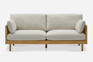 The Lucia Cane Sofa, White Wash, is a two-seater with light gray cushions, a wooden frame featuring straight legs, and two cylindrical armrest pillows on each side, all set against a plain white background.