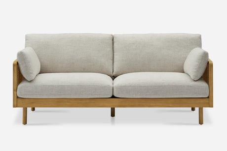 The Lucia Cane Sofa, White Wash, is a two-seater with light gray cushions, a wooden frame featuring straight legs, and two cylindrical armrest pillows on each side, all set against a plain white background.