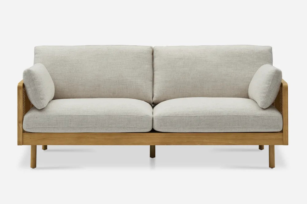 The Lucia Cane Sofa, White Wash, is a two-seater with light gray cushions, a wooden frame featuring straight legs, and two cylindrical armrest pillows on each side, all set against a plain white background.