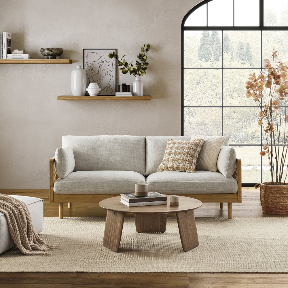 A contemporary living room featuring the Lucia Cane Sofa in White Wash, accompanied by a wooden coffee table and decorative items on a shelf. A large window offers a view of trees, while a neutral-toned rug and a potted plant add to the cozy ambiance.