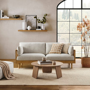 A contemporary living room featuring the Lucia Cane Sofa in White Wash, accompanied by a wooden coffee table and decorative items on a shelf. A large window offers a view of trees, while a neutral-toned rug and a potted plant add to the cozy ambiance.