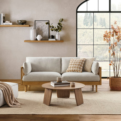 A contemporary living room featuring the Lucia Cane Sofa in White Wash, accompanied by a wooden coffee table and decorative items on a shelf. A large window offers a view of trees, while a neutral-toned rug and a potted plant add to the cozy ambiance.