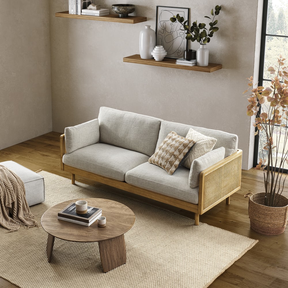 A cozy living room showcases the Lucia Cane Sofa in White Wash with patterned cushions, complemented by a round wooden coffee table adorned with books and mugs. The space is further enhanced with a beige woven rug, wall-mounted shelves displaying decorative items, and a large window that fills the room with natural light.