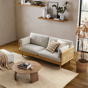 A cozy living room showcases the Lucia Cane Sofa in White Wash with patterned cushions, complemented by a round wooden coffee table adorned with books and mugs. The space is further enhanced with a beige woven rug, wall-mounted shelves displaying decorative items, and a large window that fills the room with natural light.