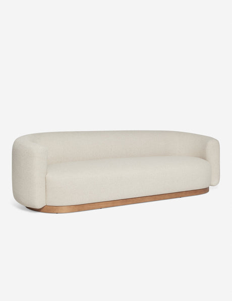 The Lowry Sofa, featuring a minimalist beige design with rounded edges and a wooden base, is showcased against a plain white background. It boasts a modern, streamlined look with cushioned armrests and a smooth, continuous back.