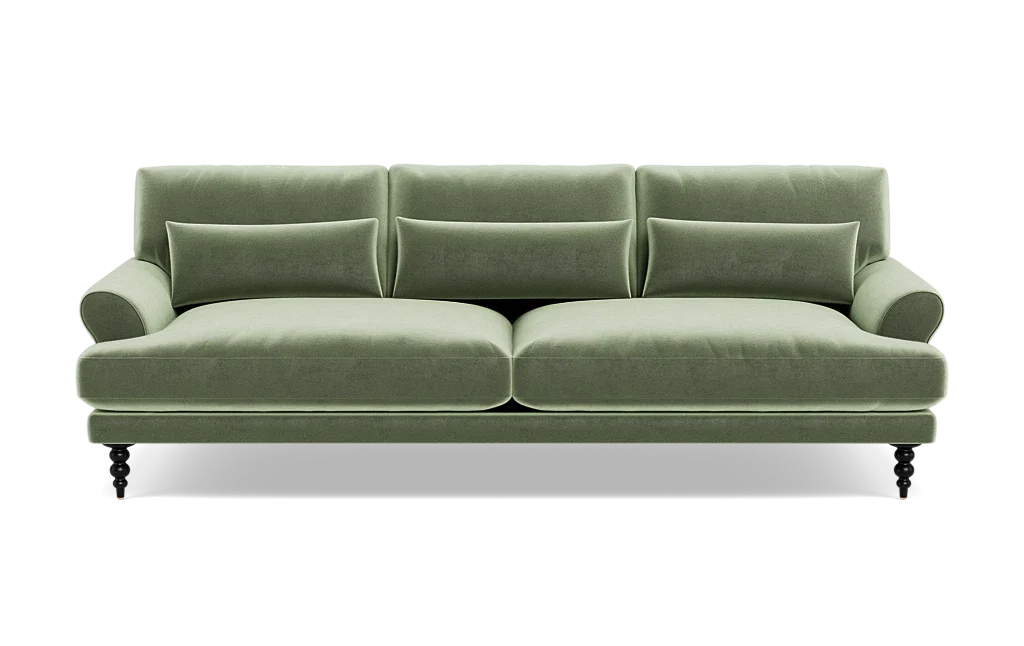 The Maxwell Custom Sofa is a green velvet piece featuring three back cushions, two seat cushions, and a pair of armrests. It comes with four small decorative pillows and rests on four dark wooden legs that give it a classic and elegant appearance.