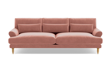 Introducing the Maxwell Custom Sofa: a charming pink velvet piece featuring three seat cushions and coordinating back cushions. It boasts elegantly rounded armrests and stands on four wooden legs adorned with intricate carvings. Enhancing its comfort are two rectangular throw pillows on the seat, all set against a simple black backdrop.