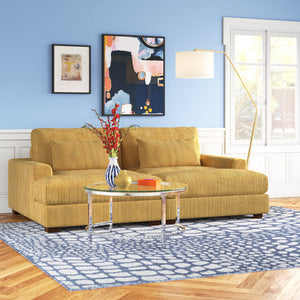 An Etta Avenue Mordecai 90'' Square Arm Sofa in yellow is placed on a blue and white patterned rug in the living room. Art is displayed on a blue wall above the sofa, and a tall floor lamp stands beside it. A glass coffee table, adorned with books and a vase of red flowers, complements the space, which is illuminated by natural light.