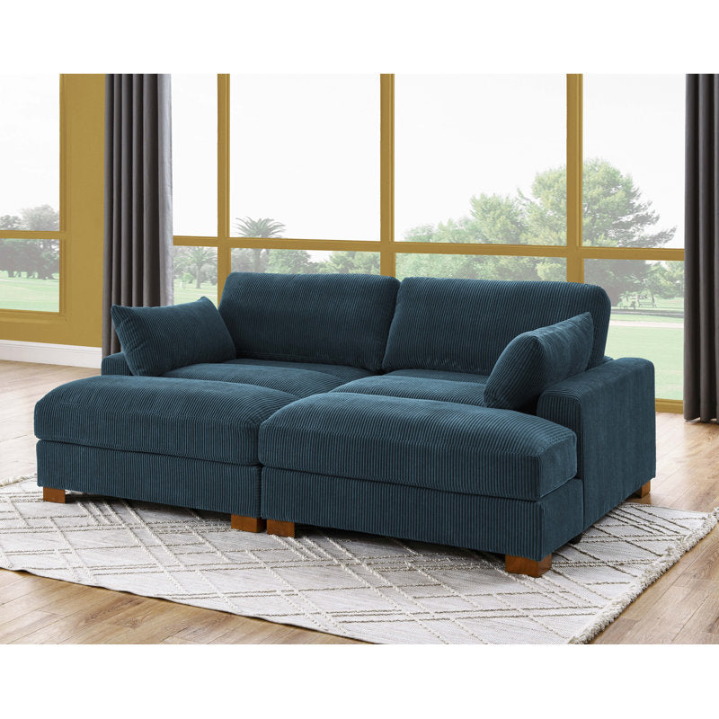 A deep teal Etta Avenue Mordecai 90'' Square Arm Sofa, made with corduroy and featuring wooden legs, is placed on a light patterned rug. The room includes large windows overlooking a lush outdoor area with trees, framed by gray curtains. The wooden floor beautifully complements the sofa and decor.