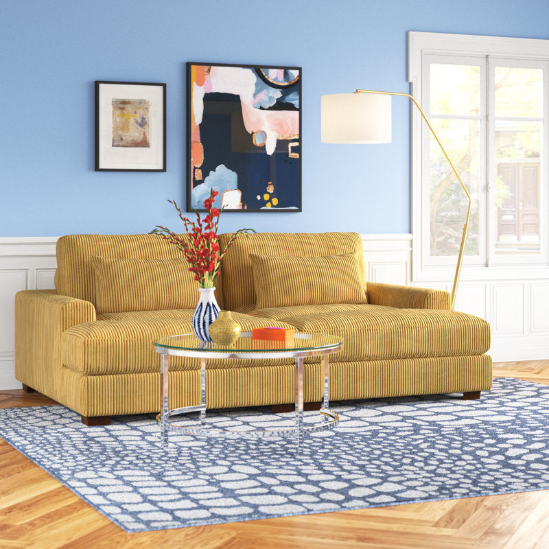 An Etta Avenue Mordecai 90'' Square Arm Sofa in yellow is placed on a blue and white patterned rug in the living room. Art is displayed on a blue wall above the sofa, and a tall floor lamp stands beside it. A glass coffee table, adorned with books and a vase of red flowers, complements the space, which is illuminated by natural light.