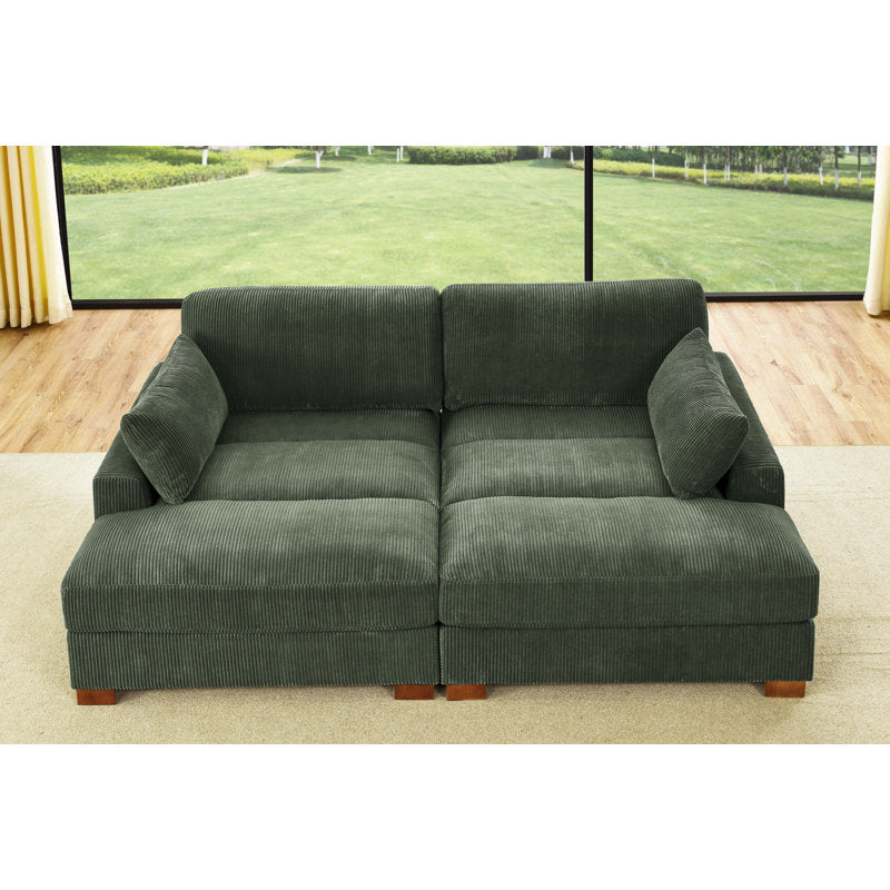 An Etta Avenue Mordecai 90'' Square Arm Sofa, upholstered in green corduroy and featuring two large cushions, is set in a bright room. Large windows in the background offer a view of a grassy outdoor area, while the floor combines light wood and carpet elements.