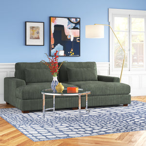 A cozy living room features the Etta Avenue Mordecai 90'' Square Arm Sofa in green corduroy, placed on a blue patterned rug. A round glass coffee table displays a vase of red flowers and books. Abstract art adorns the blue wall, while a tall floor lamp brings warmth to the space.