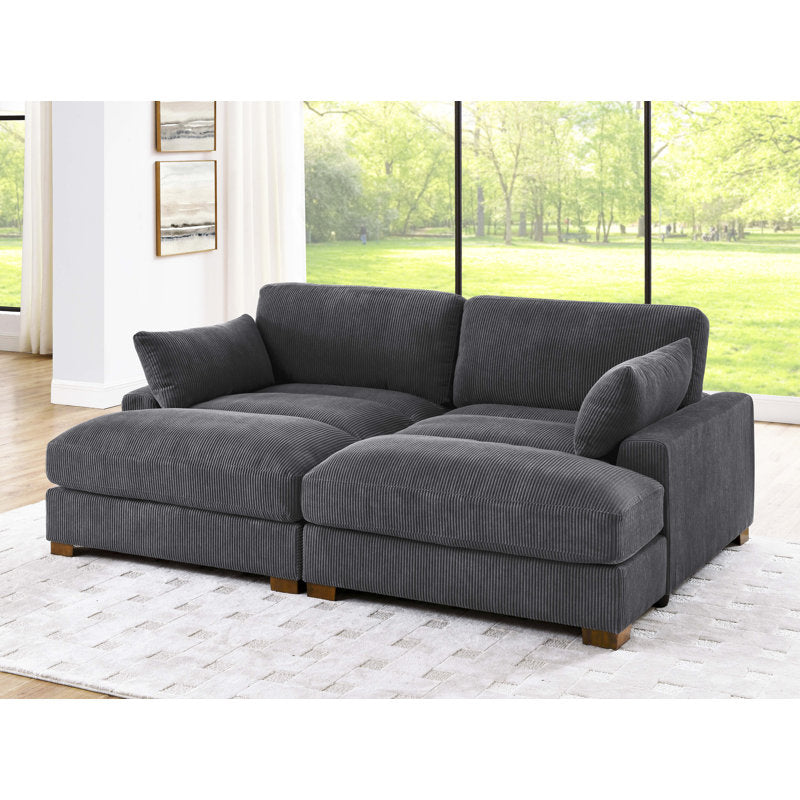 The Etta Avenue Mordecai 90'' Square Arm Sofa in dark gray, featuring plush cushions, is elegantly placed on a light-colored area rug in a bright living room. The large windows showcase a view of lush greenery outside, while framed photos on the walls enhance the cozy ambiance.