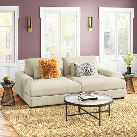 A cozy living room features the Etta Avenue Mordecai 90'' Square Arm Sofa in beige, embellished with an orange textured pillow and a patterned cushion. A round coffee table rests on a woven rug. The walls are painted mauve and have large windows accented by black wall sconces, while side tables showcase decorative items.