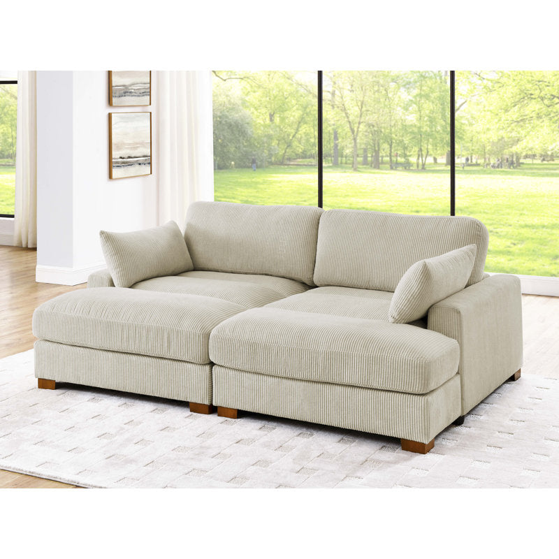 The Etta Avenue Mordecai 90'' Square Arm Sofa in beige corduroy with wooden legs is positioned on a light-colored rug in a bright room. Large windows in the background unveil a lush green outdoor scene, enhancing the cozy and inviting atmosphere.