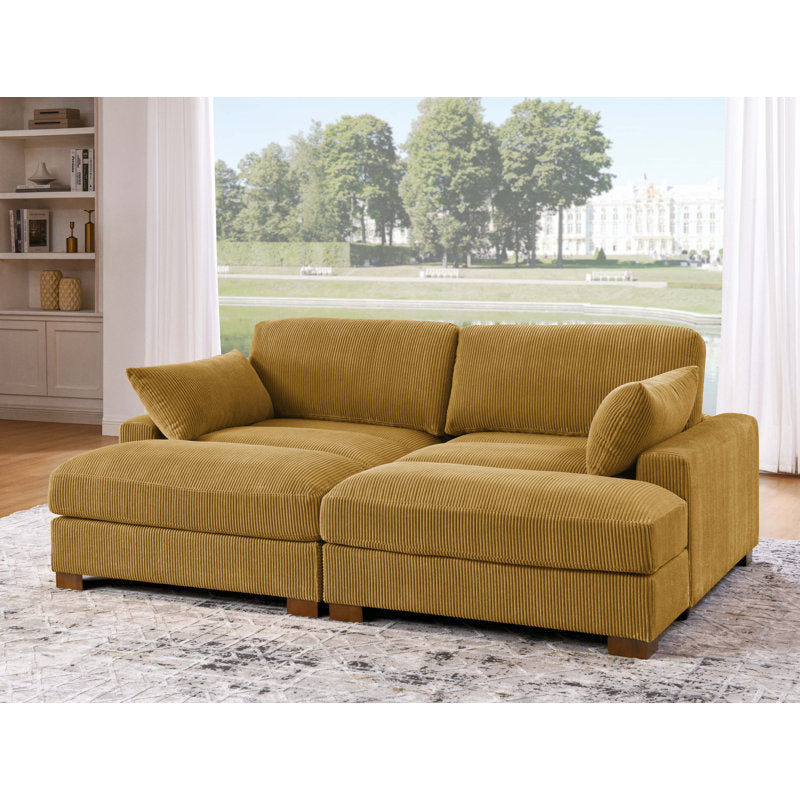 An inviting Etta Avenue Mordecai 90'' Square Arm Sofa in mustard-yellow corduroy, complete with plush cushions, graces a well-lit living room. Large windows offer a scenic view of a garden adorned with green trees and a majestic building in the distance. The floor is enhanced by the addition of a textured rug.