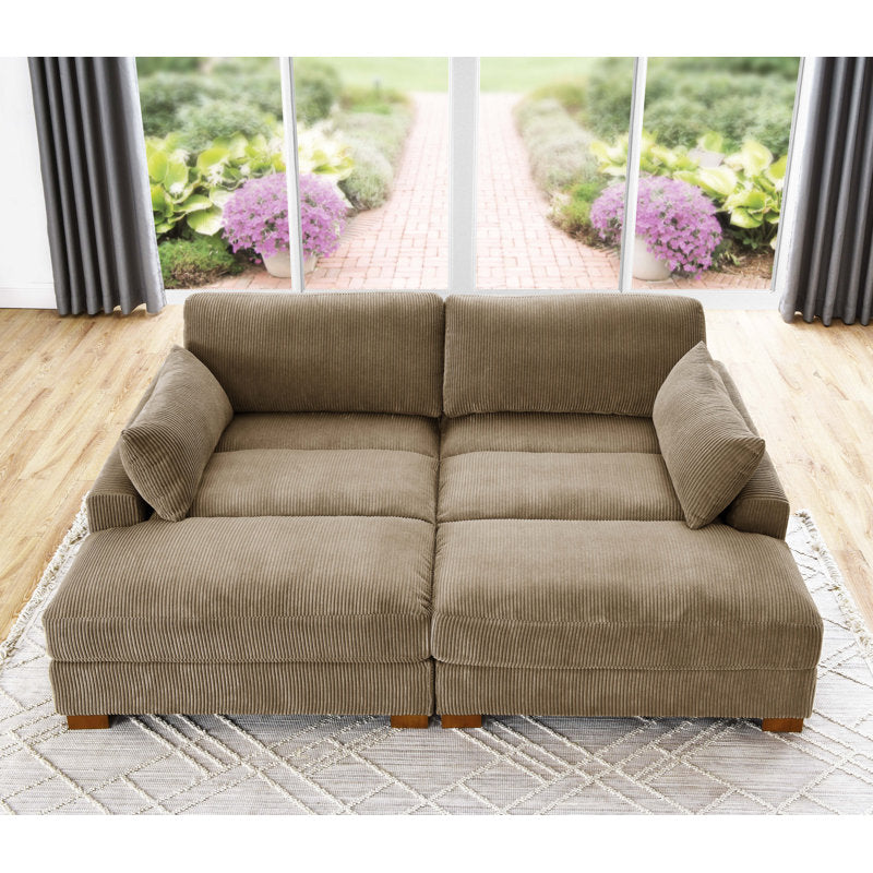 The Etta Avenue Mordecai 90'' Square Arm Sofa, a cozy brown corduroy sectional with plush cushions, is centered in a bright living room. Large windows reveal a garden path adorned with blooming pink flowers and vibrant green foliage outside. The room highlights light wood floors and a patterned rug.