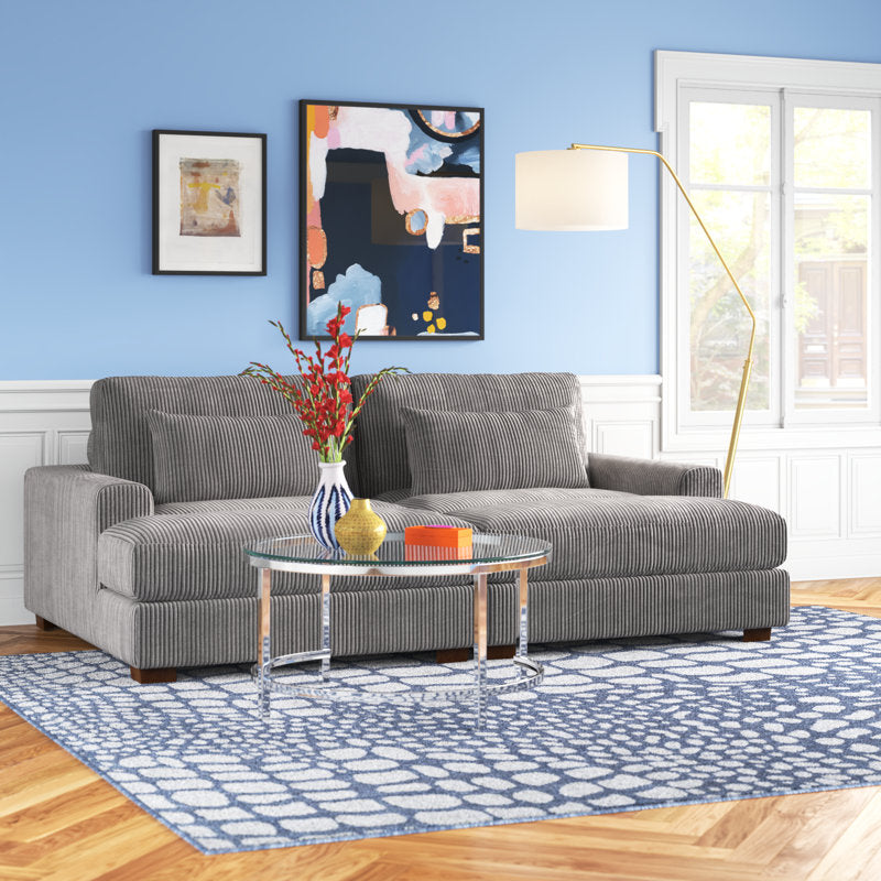 In a contemporary living room, the Etta Avenue Mordecai 90'' Square Arm Sofa in gray corduroy sits atop a blue patterned rug. In front of the sofa, a round glass coffee table displays a vase of red flowers. The walls are painted blue and adorned with abstract art, while a tall floor lamp stands beside the sofa. Large windows illuminate the space with natural light.
