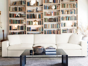 In a cozy living room, a large 4-seat modular chaise sectional with ottoman is situated in front of floor-to-ceiling bookshelves filled with assorted books. A black coffee table holds a stack of books and a rustic bowl, while soft lighting and a guitar accentuate the inviting space.
