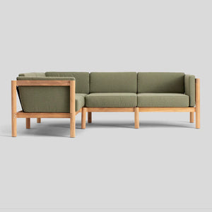 Introducing the Haven Sectional, a sleek and minimalist L-shaped sofa featuring an elegant light wooden frame and plush olive green cushions, set against a plain gray backdrop.