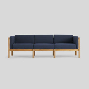The Haven Sofa is a contemporary three-seater featuring a wooden frame and dark blue cushions. Its minimalist design emphasizes clean lines and the striking contrast between the natural wood and rich blue upholstery, set against a plain light gray background.