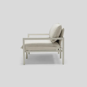 Side view of the Terra Loveseat, a modern, minimalist outdoor lounge chair featuring a light gray metal frame with plush, off-white cushions on the seat and backrest, all set against a plain gray background.
