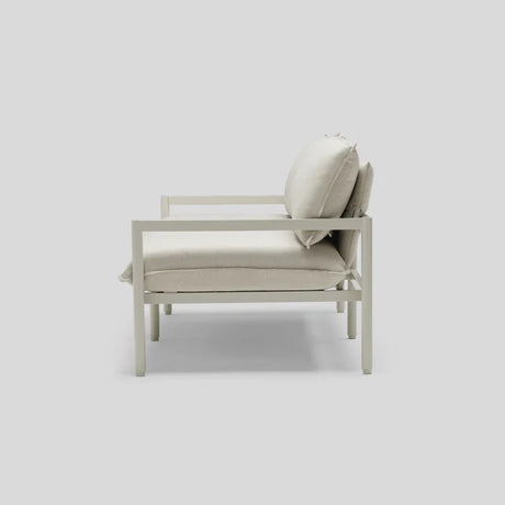Side view of the Terra Loveseat, a modern, minimalist outdoor lounge chair featuring a light gray metal frame with plush, off-white cushions on the seat and backrest, all set against a plain gray background.